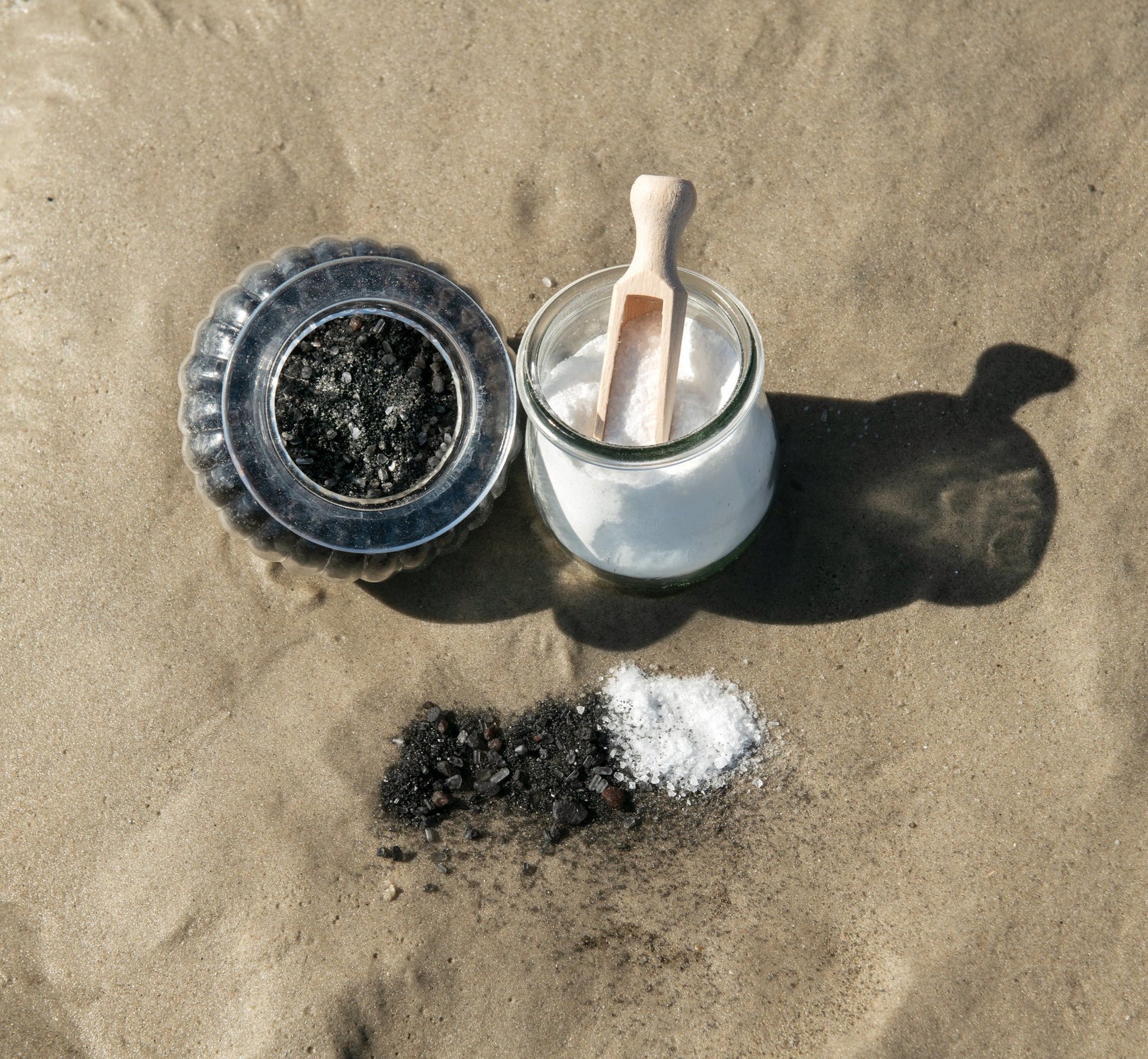 Zwei Badesalze – Basisches Aktivkohle-Bad und Alpenkräuter Vital-Bad – in Glastiegeln am Strand, naturrein, kontrastreich und pflegend für unterschiedliche Bedürfnisse von Körper und Geist